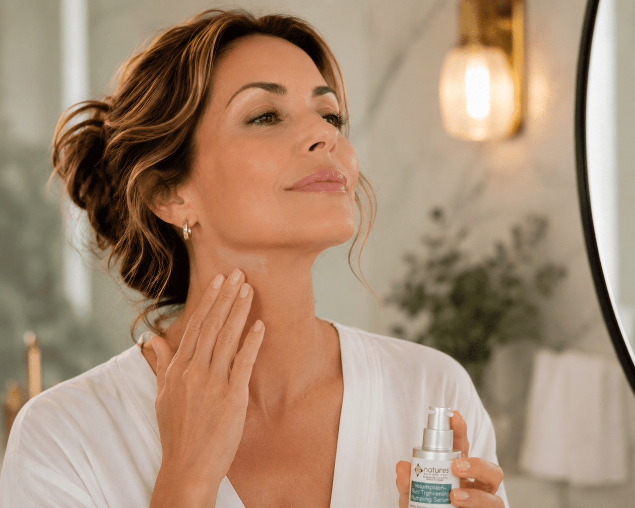 A woman in a white robe applies a skincare product from a small silver bottle to her neck, standing in front of a mirror in a softly lit bathroom.