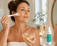 Thumbnail for A woman wrapped in a white towel applies a beige facial mask with a brush while holding a small bowl of the mask. A white bottle labeled 'Biozyme enzyme peel' with a green label sits on a bathroom counter in the background.