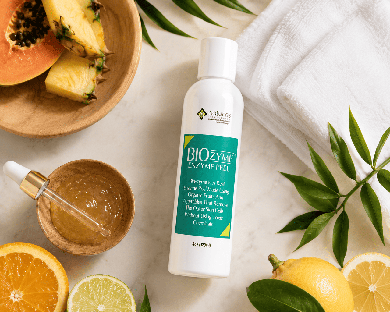 A bottle of Nature's Biozyme Enzyme Peel is displayed on a white marble surface surrounded by skincare props, including a white towel, a wooden bowl of honey, a dropper, sliced papaya, orange, lime, and pineapple.