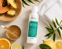 Thumbnail for A bottle of Nature's Biozyme Enzyme Peel is displayed on a white marble surface surrounded by skincare props, including a white towel, a wooden bowl of honey, a dropper, sliced papaya, orange, lime, and pineapple.