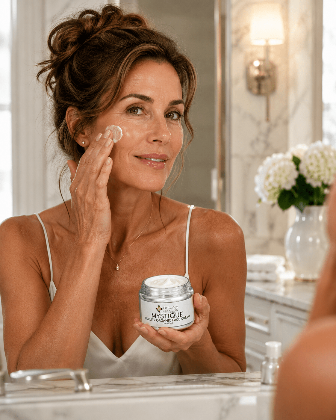 A woman in a white camisole applies cream to her cheek while holding a jar of Mystique Cream in front of a bathroom mirror.